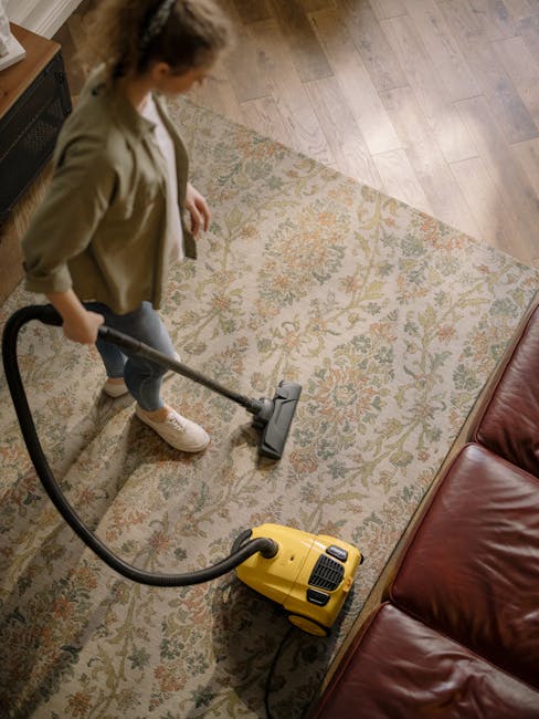 A person using a yellow vacuum cleaner to deep clean a patterned area rug with floral motifs in neutral tones, positioned on a wooden floor next to a leather sofa. The room is well-lit, and the carpet appears clean and free of dust. The individual is dressed casually, wearing a beige jacket, jeans, and white shoes, with a focus on surface cleaning in a domestic setting. This scene illustrates professional home sanitisation and deep cleaning services offered by Islington Carpet Cleaning. The background shows the edge of a wooden surface, reflecting a tidy and hygienic interior environment.