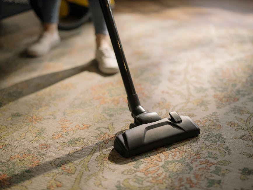 A close-up of a vacuum cleaner head actively cleaning a patterned area rug in a residential setting. The rug features a floral or traditional design with shades of beige, green, and orange. The vacuum cleaner is black with a slim nozzle, and visible in the background are the person's legs wearing dark jeans and white shoes. The scene is well-lit, highlighting the clean, dust-free surface of the rug, emphasizing surface cleaning and maintenance. This image aligns with domestic cleaning services offered by Islington Carpet Cleaning, as seen on their website regarding surface cleaning and sanitisation.