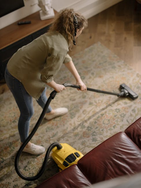 A person using a yellow vacuum cleaner to deep clean a patterned area rug with floral motifs in neutral tones, positioned on a wooden floor next to a leather sofa. The room is well-lit, and the carpet appears clean and free of dust. The individual is dressed casually, wearing a beige jacket, jeans, and white shoes, with a focus on surface cleaning in a domestic setting. This scene illustrates professional home sanitisation and deep cleaning services offered by Islington Carpet Cleaning. The background shows the edge of a wooden surface, reflecting a tidy and hygienic interior environment.