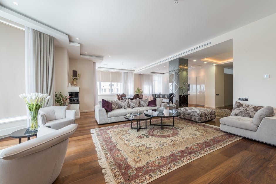 A spacious living room with light-colored walls and large windows draped with beige curtains, allowing natural light to illuminate the space. The wooden floor is covered by an ornate, traditional-styl