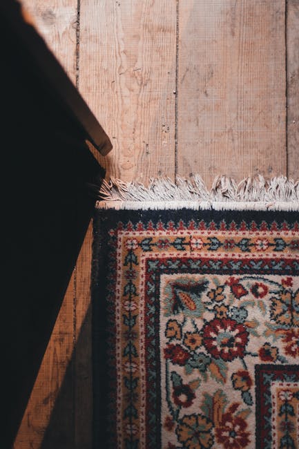 A row of colorful, patterned rugs hanging on a black metal rack outdoors along a sidewalk, with a stone brick wall behind them. The rugs display intricate designs in shades of red, orange, beige, and